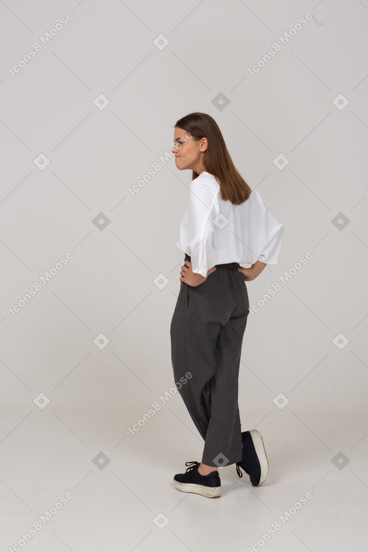 Vista posterior de tres cuartos de una joven disgustada en ropa de oficina poniendo las manos en las caderas