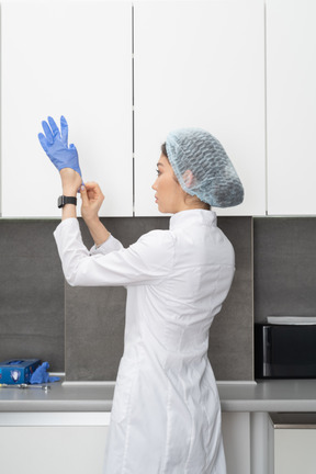 Three-quarter back view of a female nurse putting on protective gloves in her medical cabinet