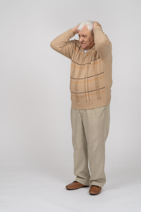 Front view of an old man in casual clothes standing with hands behind head