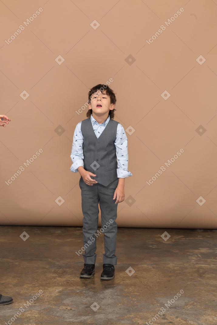 Vista frontal de um menino de terno em pé, com os olhos fechados e a boca aberta