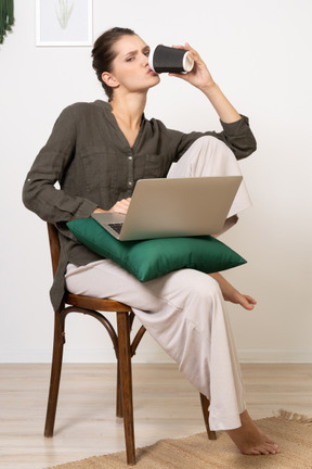 一位年轻女子坐在椅子上，拿着她的笔记本电脑和喝咖啡的前视图
