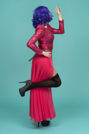 Vista lateral de uma drag queen de vestido rosa levantando uma perna e apontando para cima