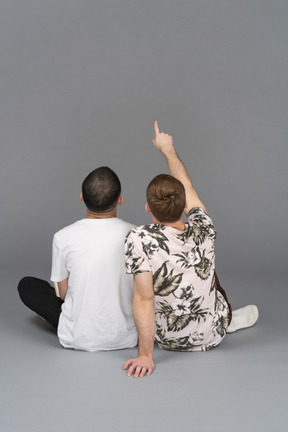 Back view of two young men sitting close to each other on the floor and pointing up
