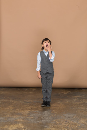 Vista frontal de un niño en traje bostezando y tapándose la boca con la mano