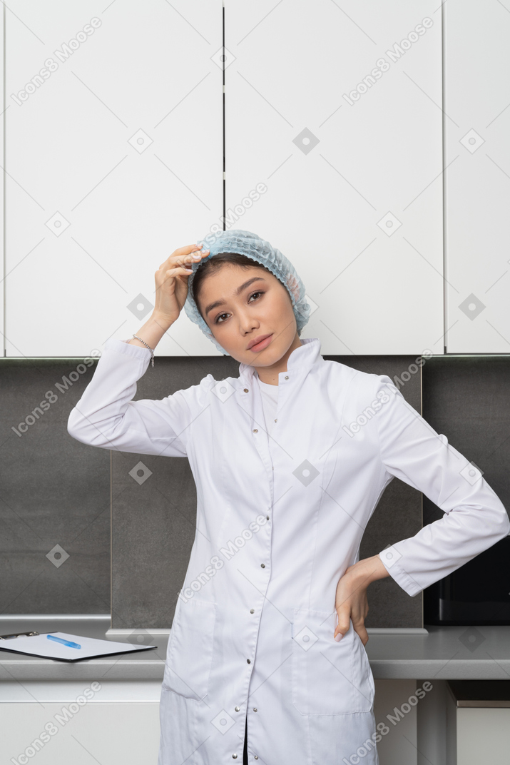 Vista frontal de una enfermera con sombrero médico tocando la cabeza y poniendo la mano en la cadera