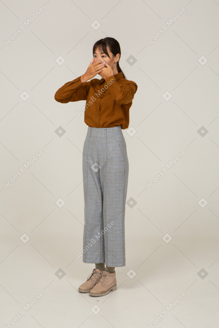 Vista di tre quarti di una giovane donna asiatica in calzoni e camicetta che nasconde la bocca