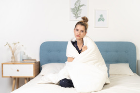 Front view of a tired young woman in pajamas wrapped in blanket staying in bed