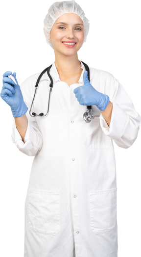 Vista frontale di una giovane dottoressa sorridente con lo stetoscopio che tiene il termometro e mostra il pollice in su