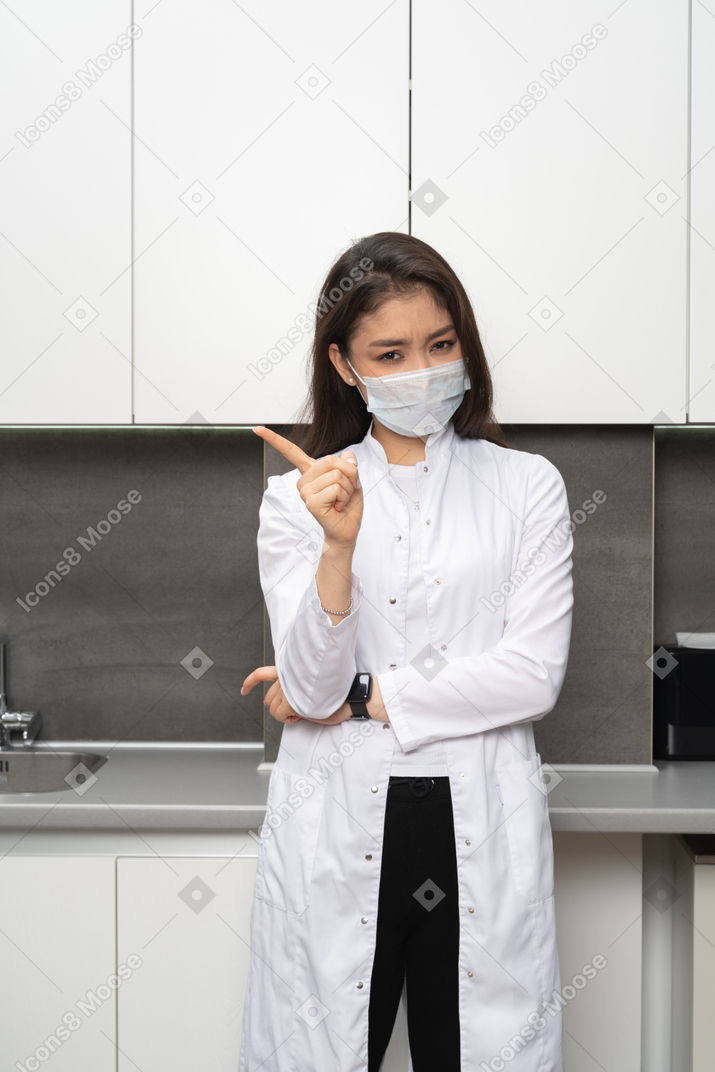 Vista frontale di una dottoressa seria in una maschera che punta il dito e che guarda l'obbiettivo
