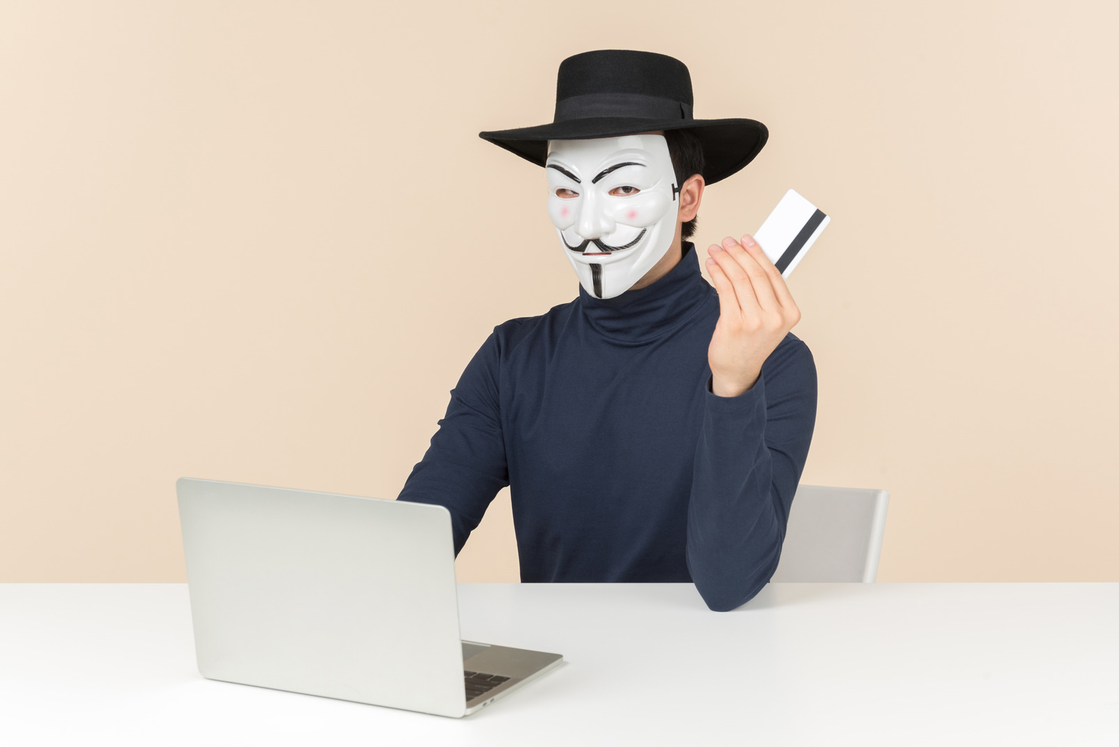 Hacker wearing vendetta mask sitting at the laptop and holding bak card