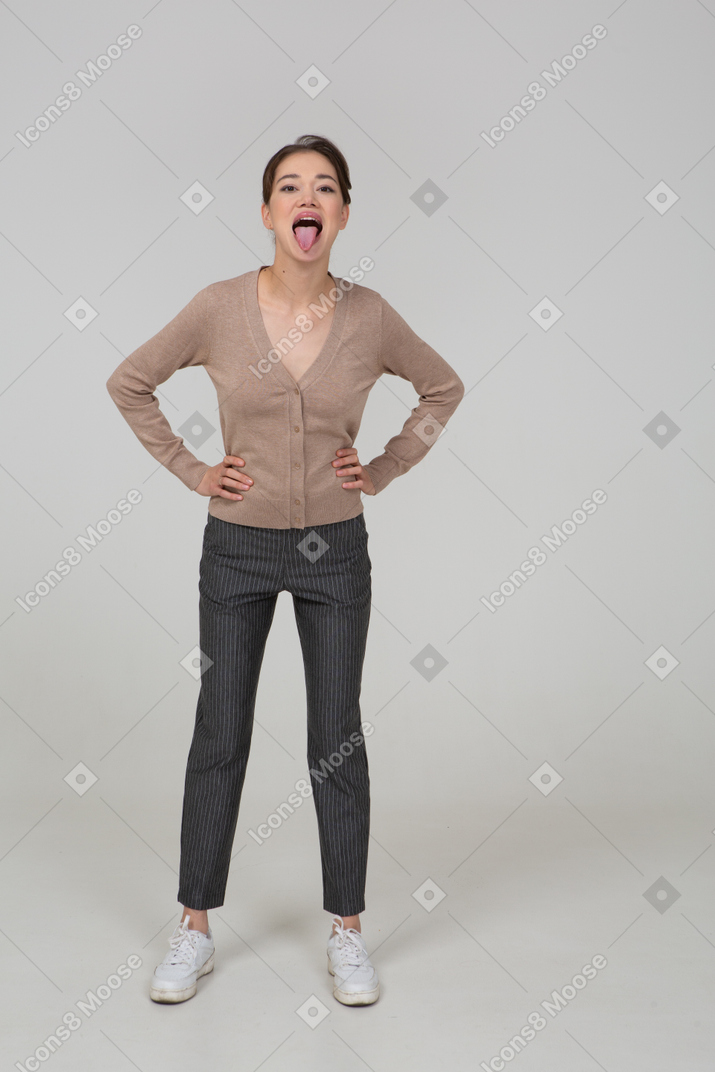 Vue de face d'une jeune femme en pull et pantalon mettant les mains sur les hanches et montrant la langue