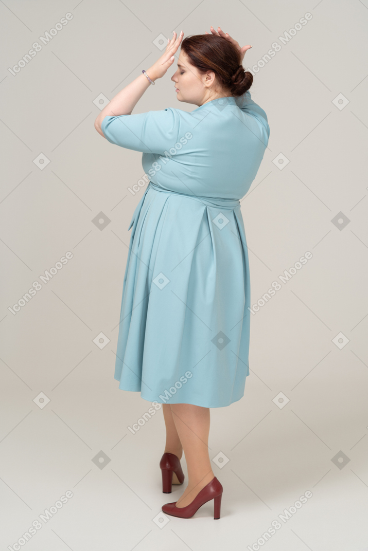 Вид сзади женщины в голубом платье, стоящей с руками на голове