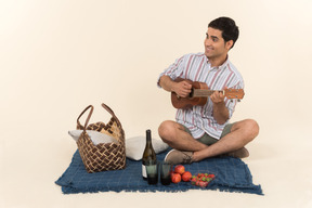 Young caucasian guy sitting on blanket and playing small guitar