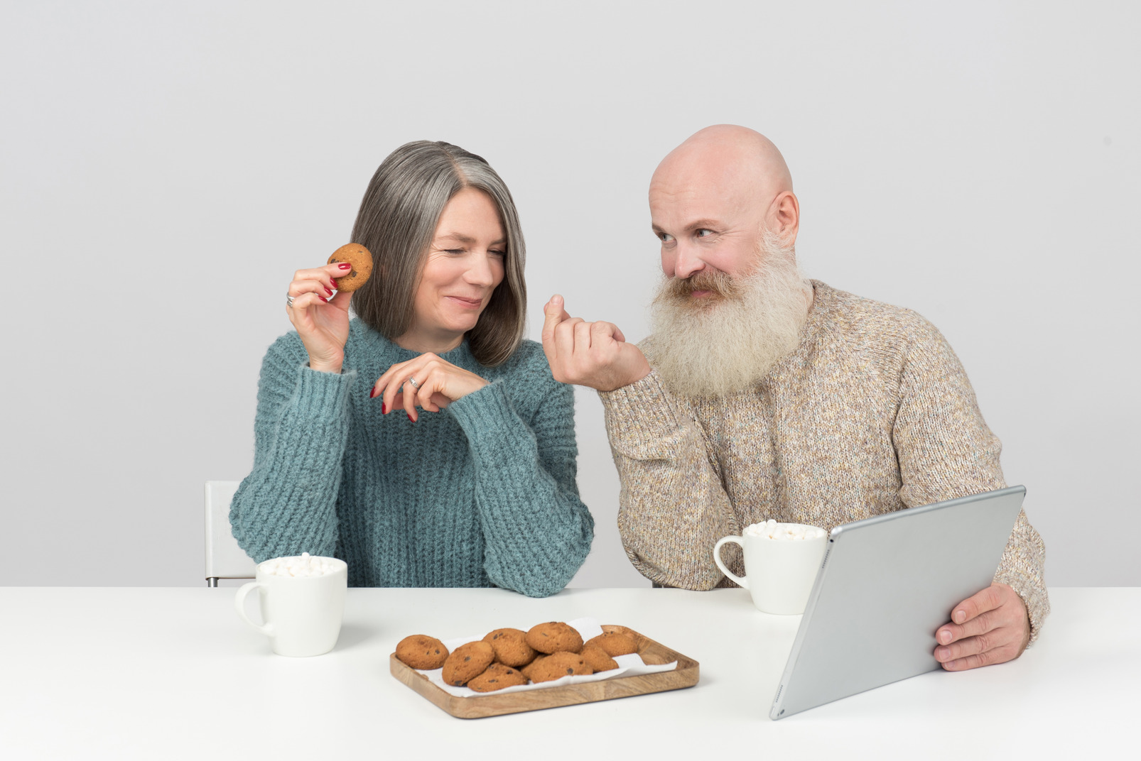 Aged couple sitting at the table, having cookies and coffee and watching movie on tablet
