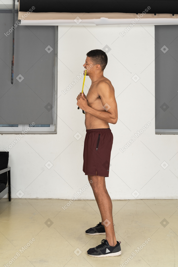 Standing in profile young man in sport shorts biting jumping rope
