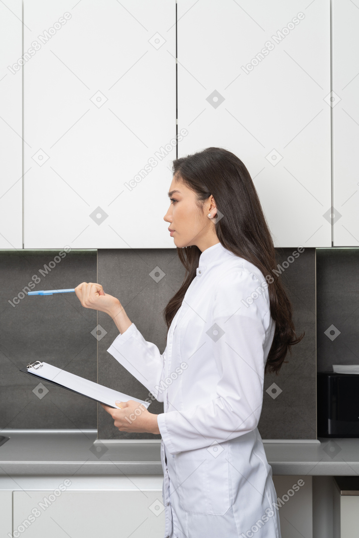 Side view of a questioning female doctor looking aside and holding a pen with a tablet