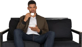 Front view of a young dreaming man sitting on a sofa while drinking coffee