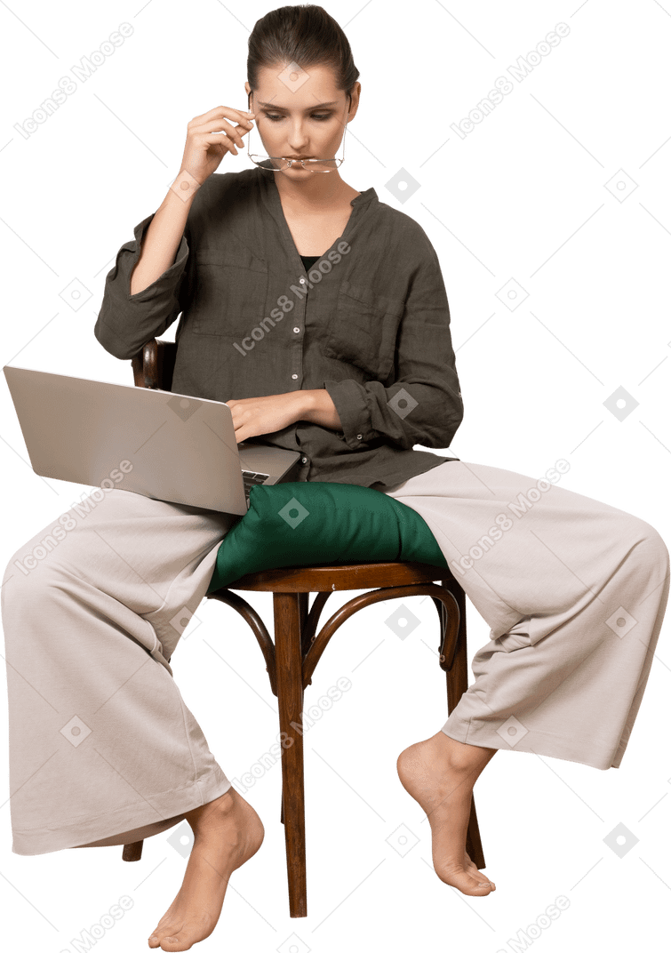 Front view of a young woman wearing home clothes sitting on a chair with a laptop and putting on glasses