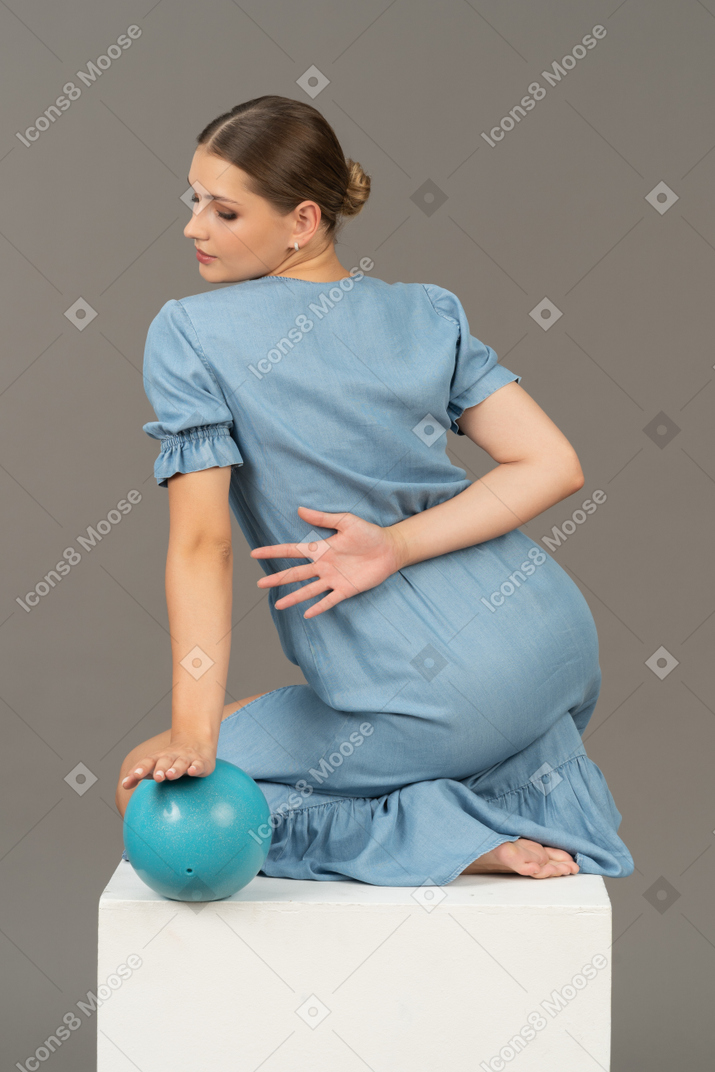 青いボールと立方体に座っている若い女性の4分の3の背面図
