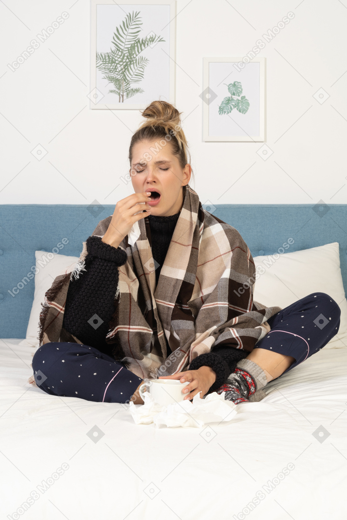 Vista frontal de una joven enferma bostezo en pijama envuelto en una manta marcada en la cama