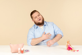Rire grand homme assis à la table avec des produits cosmétiques et poupée barbie sur elle