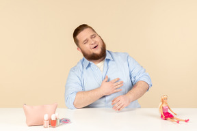 Laughing big man sitting at the table with cosmetics and barbie doll on it