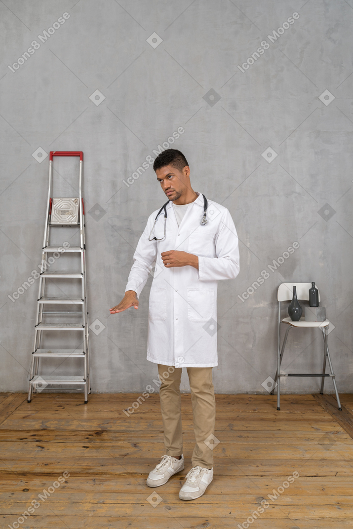 Vista de tres cuartos de un joven médico de pie en una habitación con escalera y silla que muestra el tamaño de algo