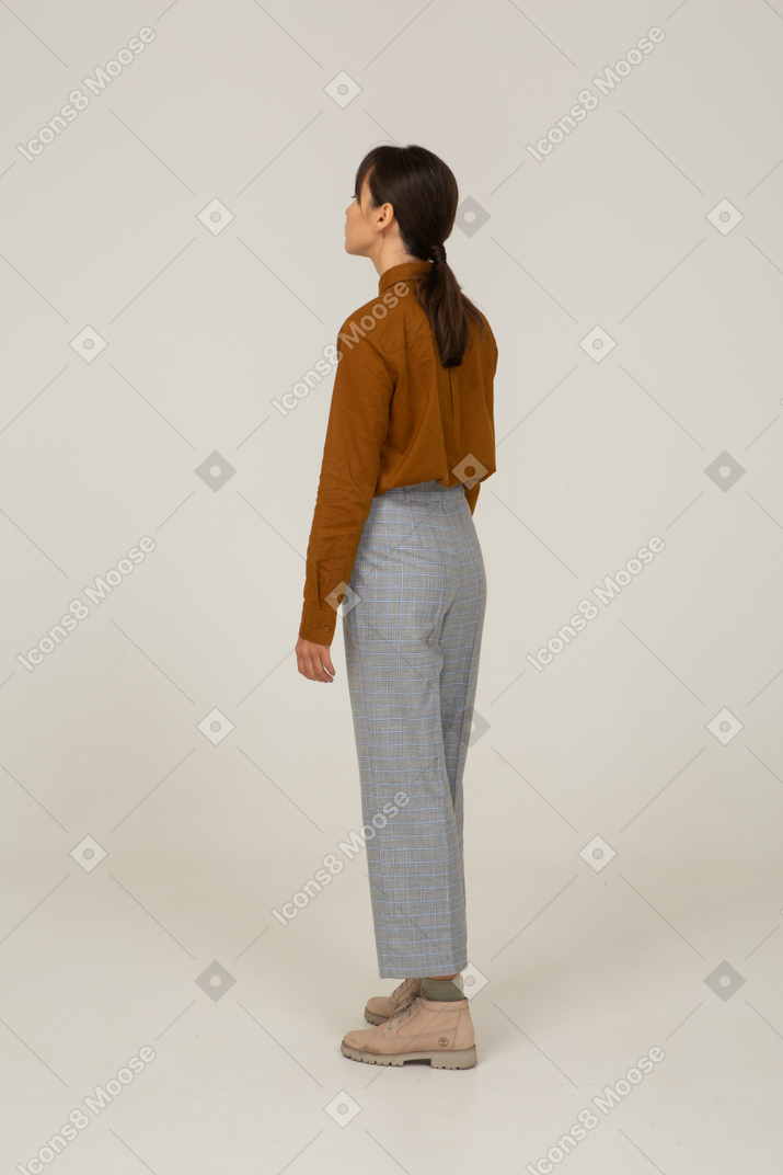Вид сзади в три четверти молодой азиатской женщины в бриджах и блузке, стоящей на месте