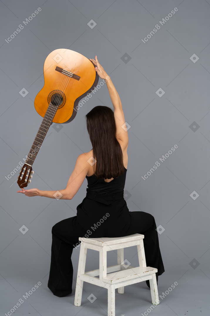 Back view of a young lady in black suit holding the guitar over head and sitting on stool