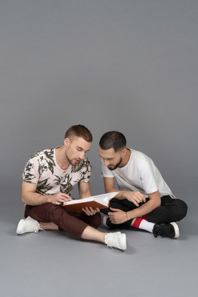Vista frontal de dos jóvenes sentados en el suelo y estudiando un libro