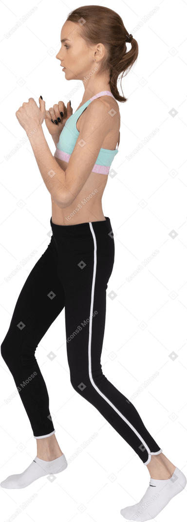 Vista lateral de una jovencita en ropa deportiva dando un paso adelante mientras aprieta los puños