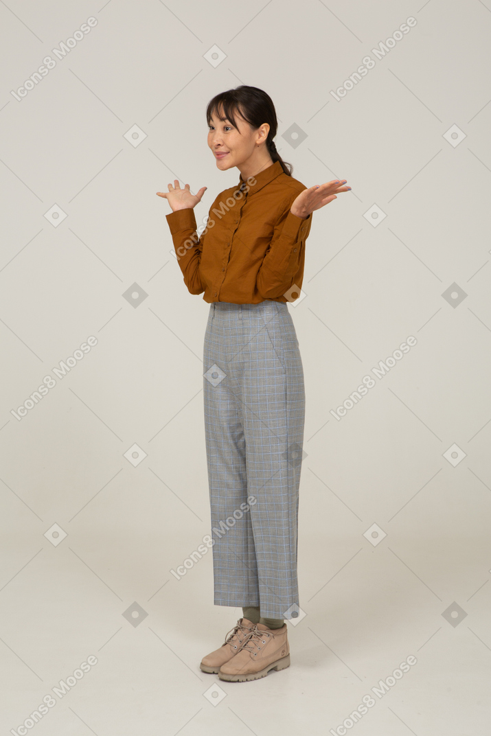 Vista di tre quarti di una giovane donna asiatica felice in calzoni e camicetta che alza le mani