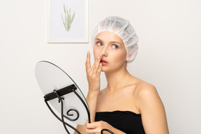 Frau in chirurgischer kappe, die spiegel hält und ihre nase berührt