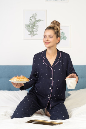 Front view of a young lady in pajamas holding a cup of coffee and some pastries sitting in bed