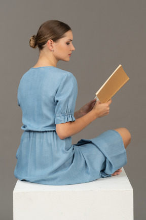 Vista lateral de uma jovem sentada em um cubo e lendo livro