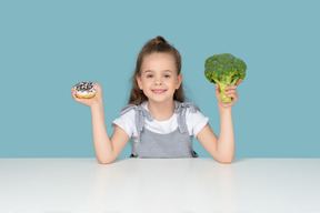 Menina bonitinha tentando escolher entre um donut e alguns brócolis