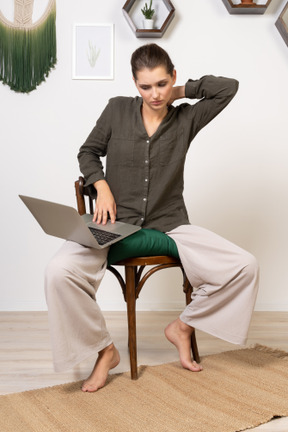 Front view of a tired young woman wearing home clothes sitting on a chair with a laptop