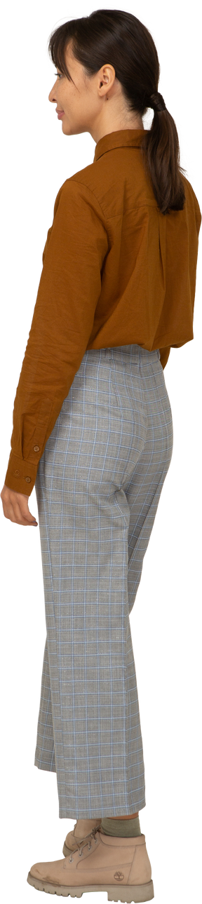 Vista posteriore di tre quarti di una giovane donna asiatica imbronciata in calzoni e camicetta in piedi ancora