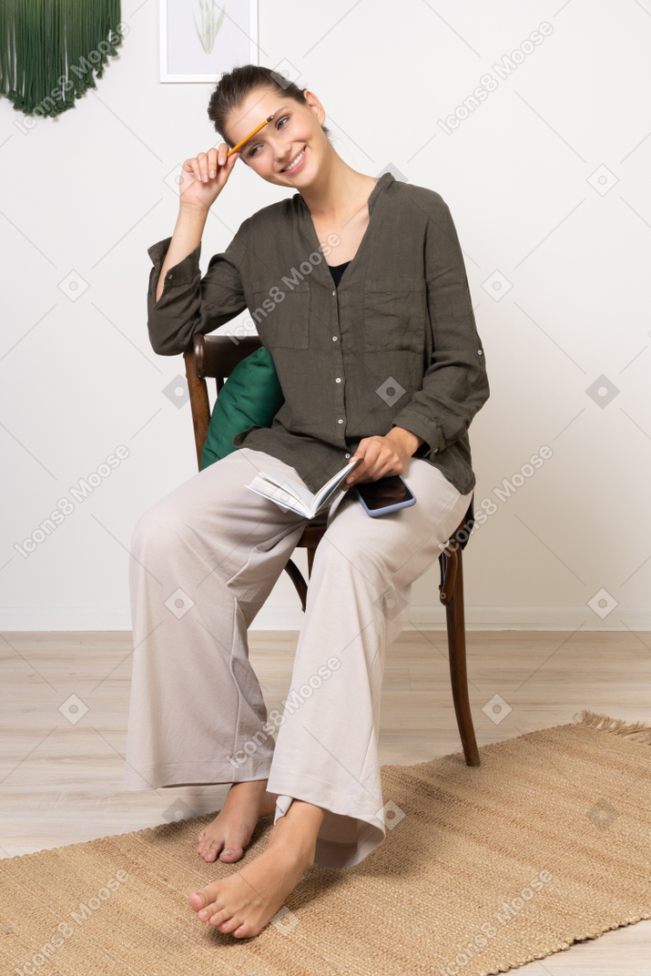 Front view of a smiling young woman wearing home clothes sitting on a chair with pencil and notebook