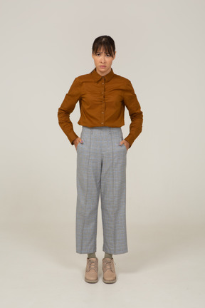 Vista frontal de una joven mujer asiática en pantalones y blusa poniendo las manos en los bolsillos