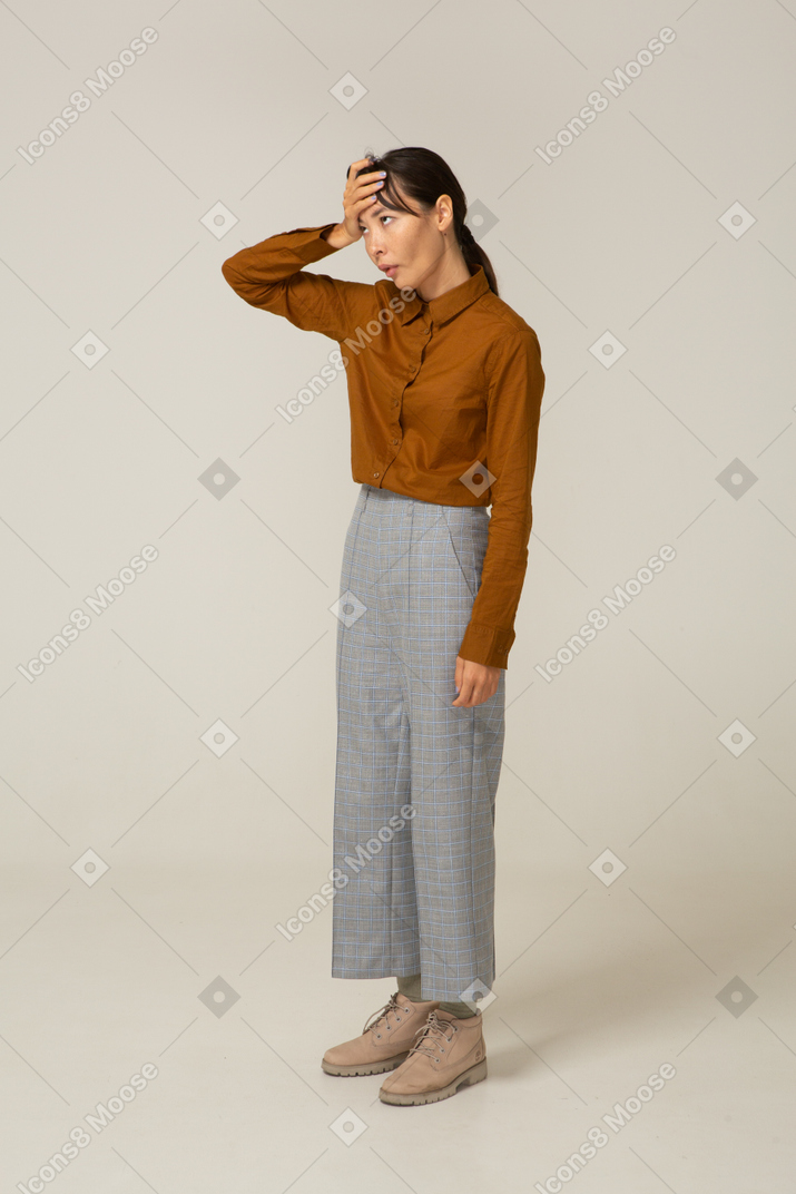 Vista de três quartos de uma jovem asiática cansada de calça e blusa tocando a cabeça