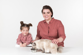 Mãe e sua filha, vestindo roupas vermelhas e cor de rosa, sentado à mesa de jantar com seu gato de família