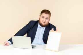 Young overweight man sitting at the desk and holding white picture frame