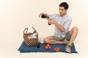 Young caucasian man sitting on the blanket and pouring wine into glass