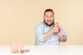 Sonriente tímido joven grande sentado a la mesa y sosteniendo la muñeca barbie