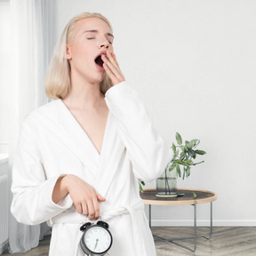 A woman in a white bathrobe is waking up with an alarm clock