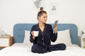 Vor einer jungen frau im pyjama, die mit der tasse kaffee im bett sitzt, während sie instagram überprüft