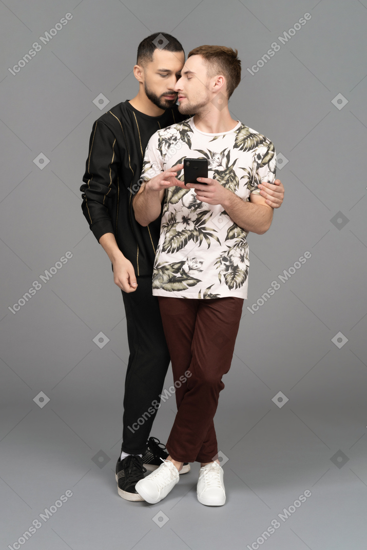 Young man half-hugging young man with the phone sensually from the back