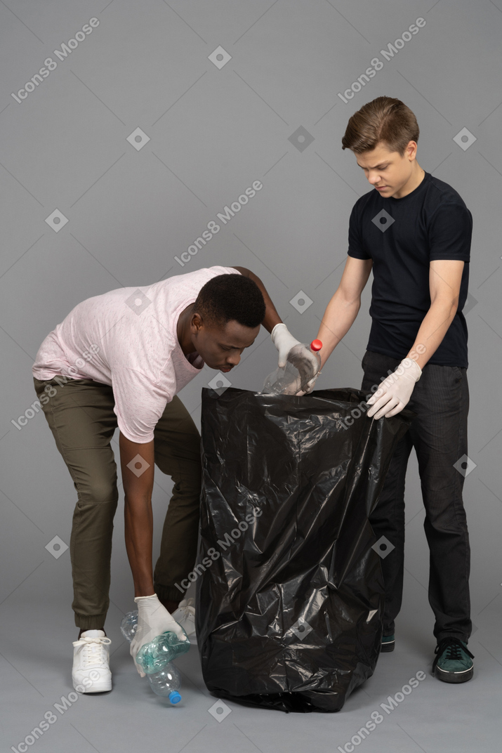 Jeune homme aidant son ami à mettre des bouteilles en plastique dans un sac poubelle