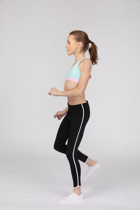 Vista lateral de uma adolescente em roupas esportivas inclinando os ombros e levantando a perna